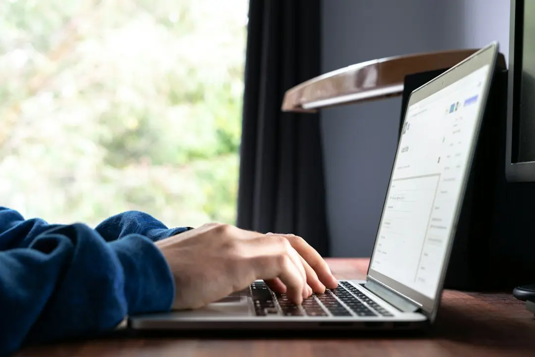 A person typing on a laptop at a desk by a window while comparing IPTV services