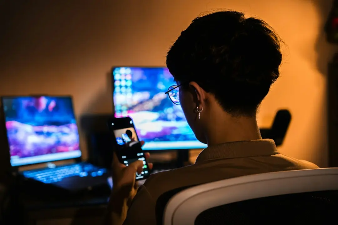 A person checking a phone in front of a laptop and monitor in a warmly lit room